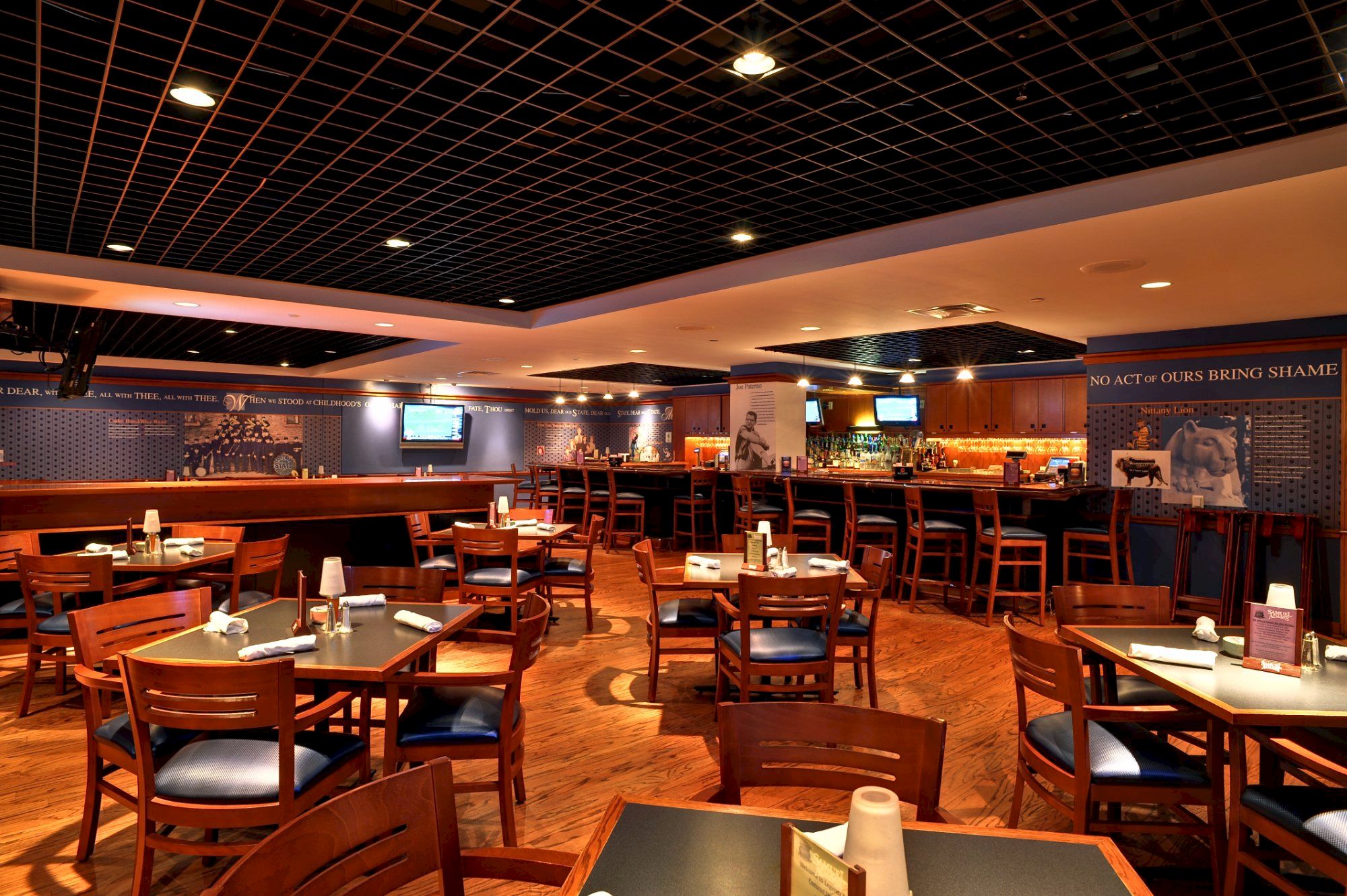 A spacious restaurant with wooden tables and chairs, dim lighting, and a bar area in the back; empty tables ready for guests.