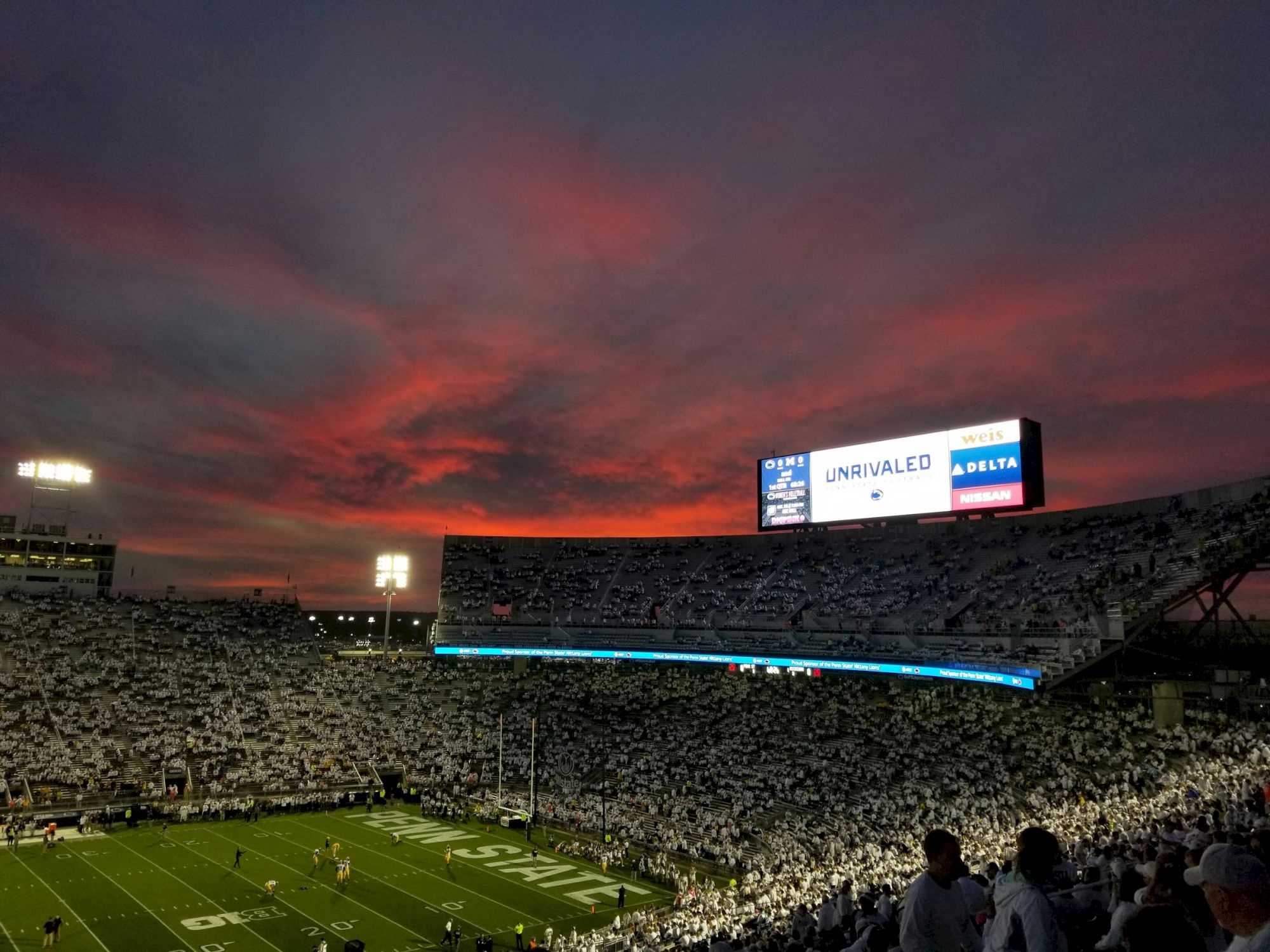 A crowded football stadium at dusk with a glowing sunset, players on the field, and a large scoreboard in the distance.