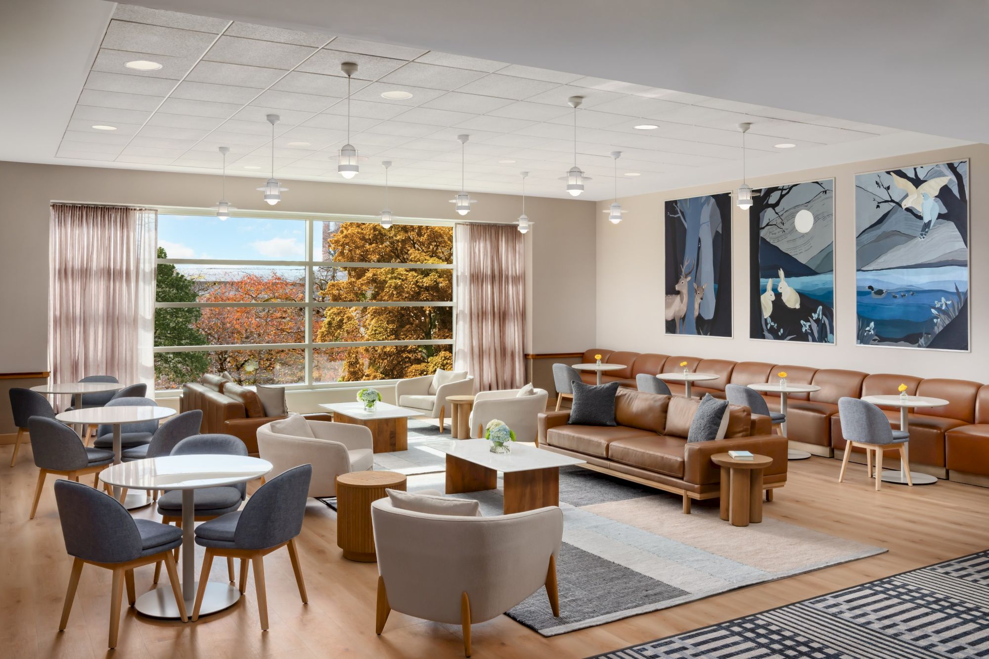 A spacious, modern lounge with multiple seating areas, warm wood floors, large windows, and contemporary art above a brown leather sofa.