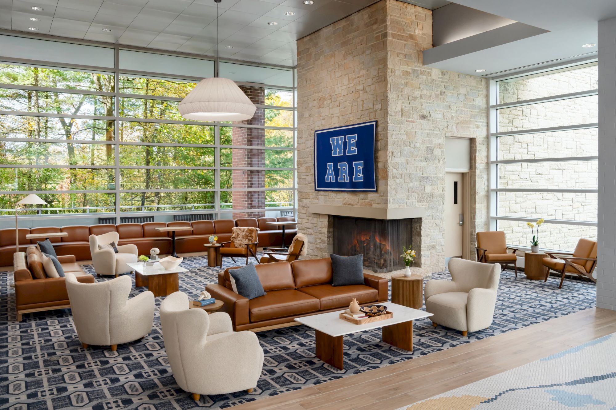 A modern lounge with cream armchairs, brown leather sofas, wooden coffee tables, large windows, a stone fireplace, and a patterned blue rug, creating a bright, airy lobby.
