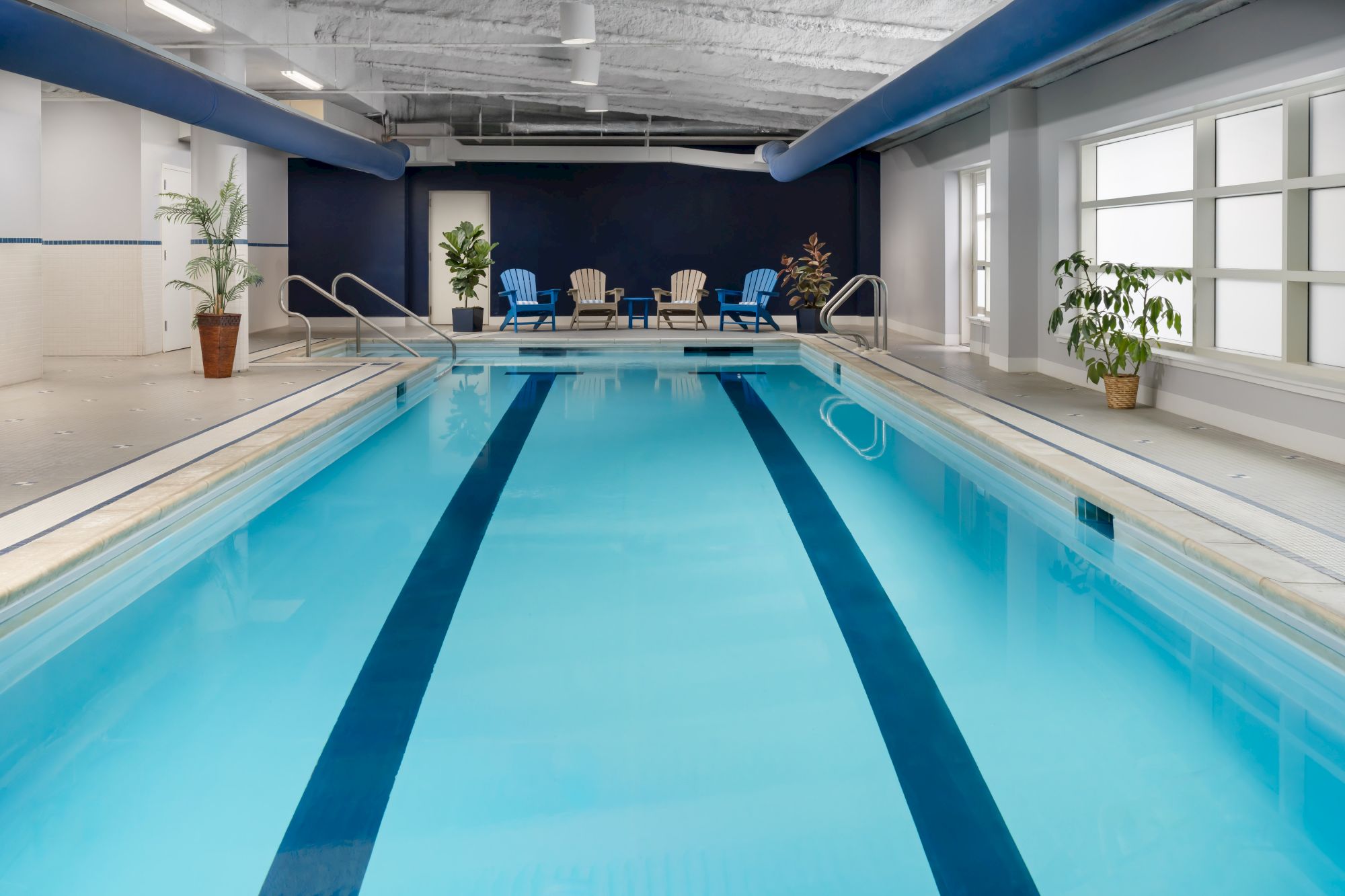 Indoor swimming pool with bright blue water, lanes, tiled deck, and lounge chairs at the far end.