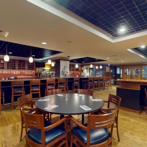 A quiet, empty bar with a long wooden counter, high stools, round tables, and warm lighting in a spacious pub-like setting.