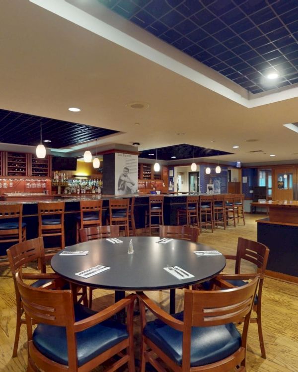 A quiet, empty bar with a long wooden counter, high stools, round tables, and warm lighting in a spacious pub-like setting.