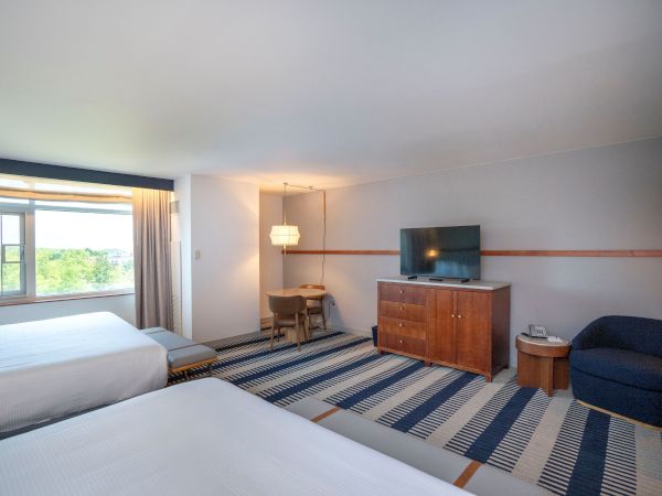 Twin beds, a desk with chair, a dresser with a wall-mounted TV, a small sofa, and a striped blue rug in a bright hotel room with a window view, 140 characters exactly, ending with a period.