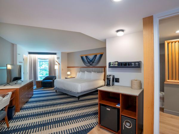 A hotel room with a king bed, desk area, flat-screen TV, blue patterned carpet, seating nook by the window, mini-fridge, and a small bathroom door to the right, all neatly arranged.