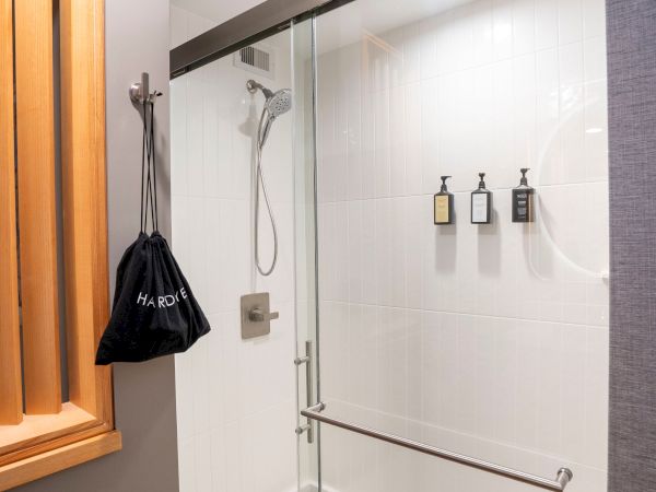 A glass shower enclosure with a chrome handle, white tiled walls, mounted shampoo bottles, a handheld shower, and a black bag hanging on a wooden cabinet, end.