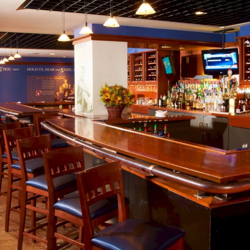 A well-lit bar with wooden counter, stools, shelves of liquor, taps, and TVs, inviting patrons for drinks in a cozy, upscale setting.