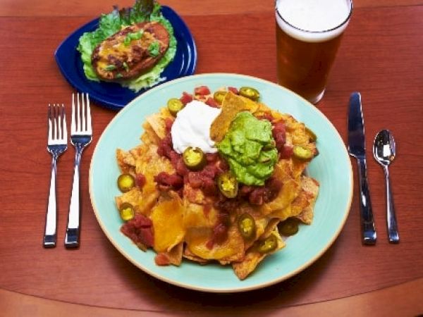 Nachos with melted cheese, beans, corn, salsa, guacamole, sour cream on a turquoise plate; side salad and a glass of beer, at a wooden table.
