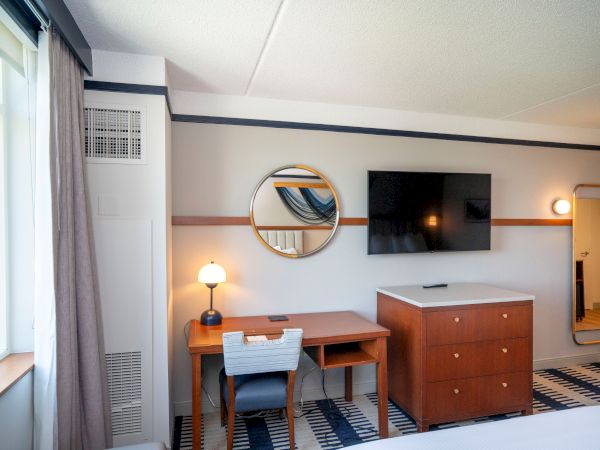 A tidy hotel room with a desk, chair, round wall mirror, wall-mounted TV, wooden dresser, full-length mirror, patterned rug, window with curtains, and soft lighting.