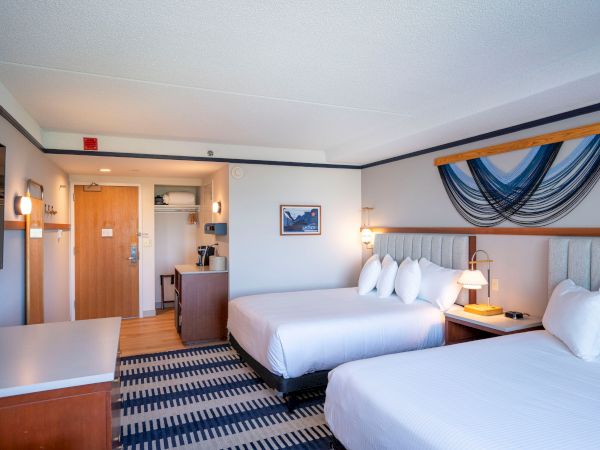 A hotel room with two double beds, bedside lamps, a chair, desk, and a decorative blue wall hanging above the headboards, near a kitchenette area.