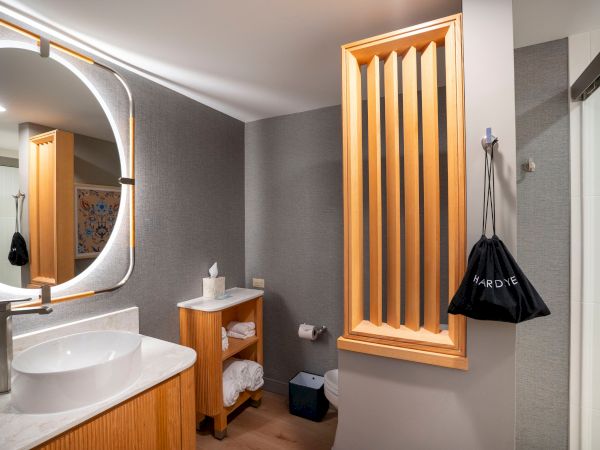 A modern bathroom with a round illuminated mirror, white sink on wood vanity, wooden shelving, and a tall vertical wooden cabinet with slats, plus a black bag hanging near the shower.