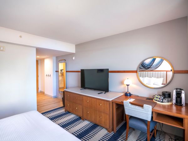 Hotel room with a bed, wooden dresser and TV, a desk with a mirror, and a patterned blue rug; bright, modern and tidy.