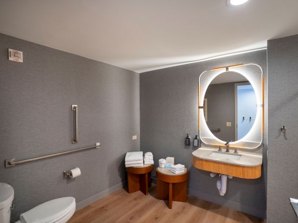 A modern bathroom with gray walls, a rounded backlit mirror above a wooden vanity, two small stools with towels, a toilet, and accessible grab bars.