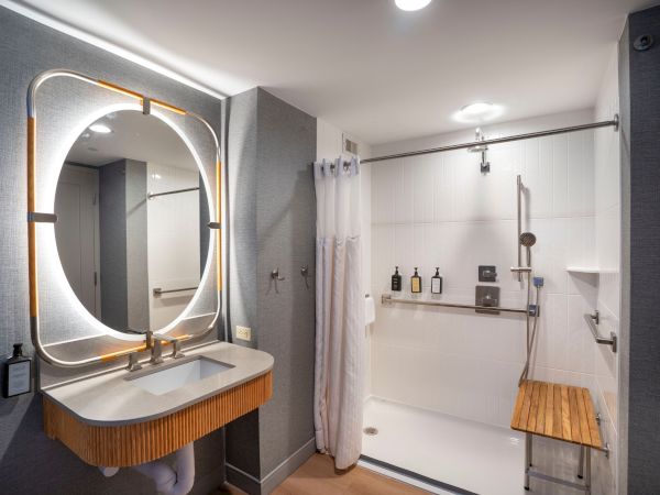 A modern bathroom with a rounded backlit mirror above a wooden vanity, and a glass-enclosed shower with a wooden bench inside.