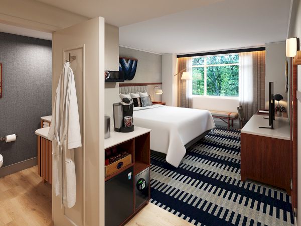 A hotel room with a king bed, desk, dresser, and large window; a small bathroom area with a towel hanging, and a striped carpet running through the room.