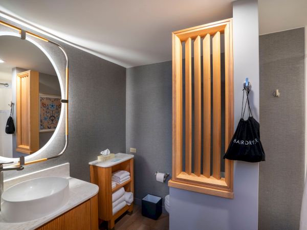 A modern bathroom with a round backlit mirror, white basin on a wooden vanity, open shelf with towels, and a wooden slatted wall feature by the doorway, plus a hanging bag and neutral tones.