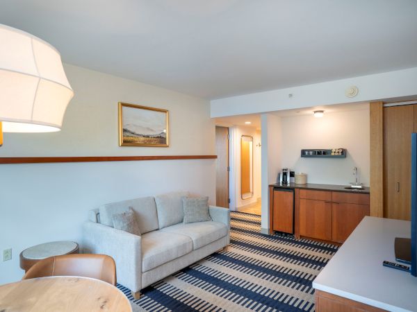 A modern hotel suite living area with a light grey sofa, striped carpet, wall art, a round pendant light, and a kitchenette with coffee station; entry hallway ahead.