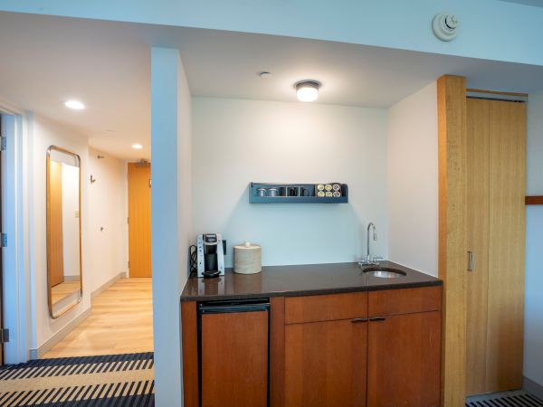 A compact kitchenette with wooden cabinets, a small sink, a coffee maker, a microwave, and a countertop—neatly arranged in a hotel-like suite.