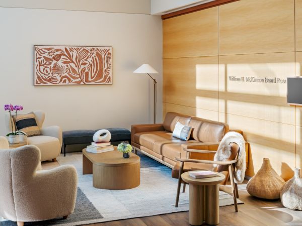 A modern, cozy living room with tan leather sofa, curved chairs, round coffee tables, a textured rug, plants, art on the wall, and warm wood paneling.