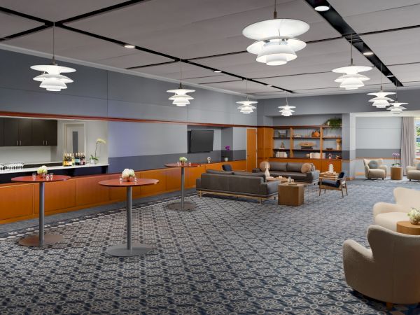 A modern lounge with orange and gray accents, multiple sofas, low coffee tables, a patterned carpet, and pendant ceiling lights in a spacious, airy setting.