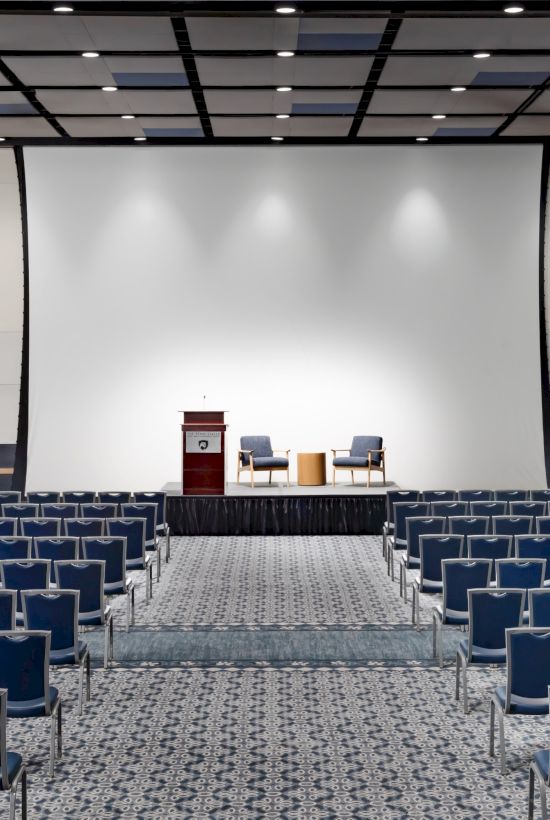 A large conference hall with a stage, podium, and rows of blue chairs facing a big projector screen.