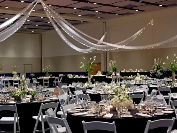 A banquet hall set for a formal event, round tables with black tablecloths, white chairs, elegant floral centerpieces, and ceiling drapery with soft lighting.