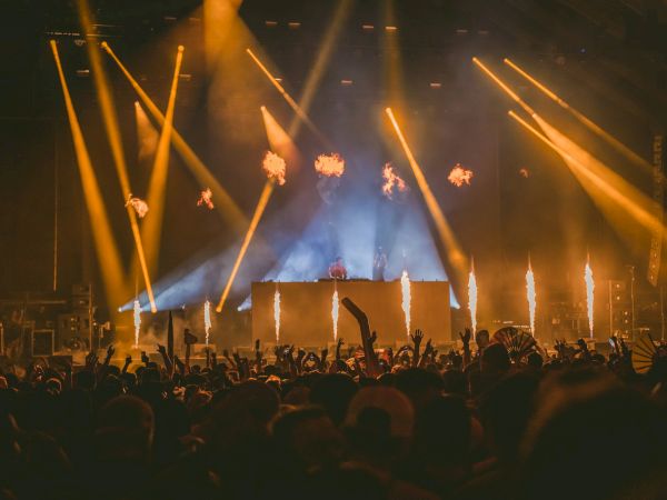 A large crowd at a concert with a DJ booth, vibrant orange stage lights, and pyrotechnics lighting the air, creating an energetic nightlife vibe.