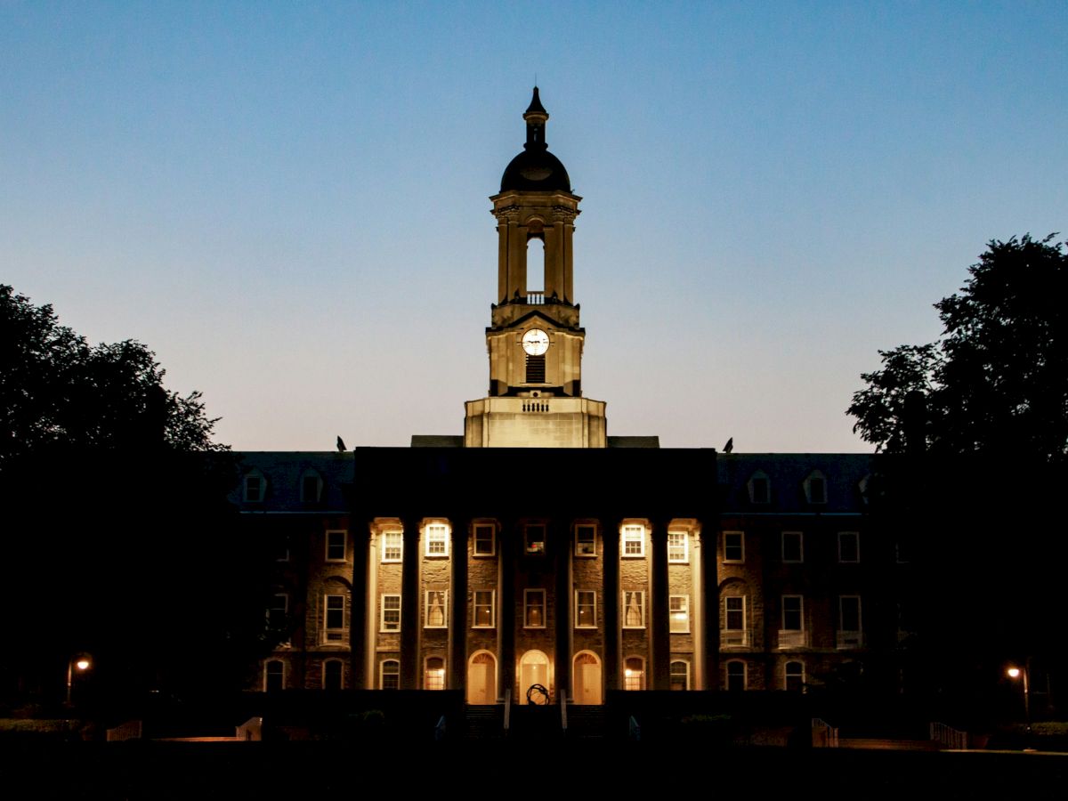 A grand, historic university building lit at dusk, with a central clock tower and columns, framed by trees on either side.