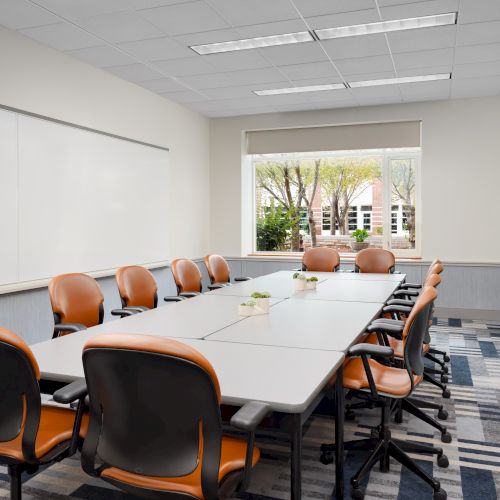 A modern conference room with a long table, orange and black chairs, a large whiteboard, and a window view outside, ready for a meeting.