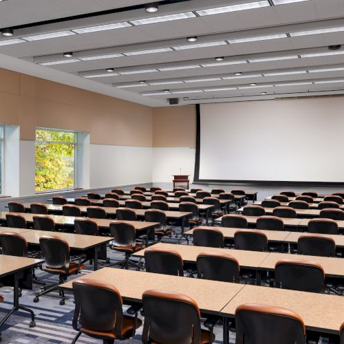 A large, empty college lecture hall with rows of desks and chairs facing a projection screen and podium, windows on the side, bright and neat.