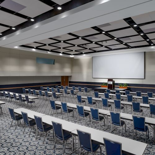 A large conference room with rows of blue chairs and white desks facing a stage and a big projection screen, set for a presentation.