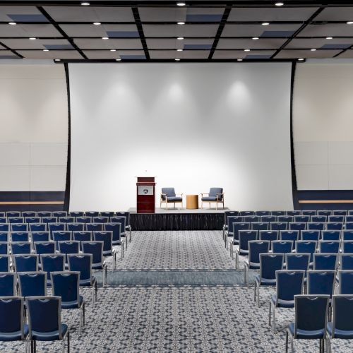 An empty conference hall with a stage, podium, two chairs, and rows of blue chairs facing a large white screen, ready for a presentation.