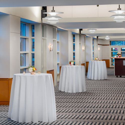 A bright event space with high windows, round cocktail tables draped in white cloth, and soft ceiling lights along a patterned carpet.