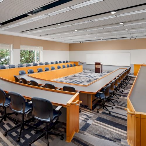 A large modern conference room with a U-shaped desk, rows of blue chairs, a whiteboard, podium, and natural light from windows, set for a formal meeting.