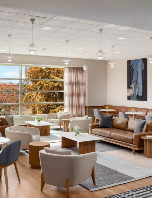 A modern, airy lounge with tan sofas, gray chairs, round tables, and large windows letting in natural light; calming wall art completes the space.